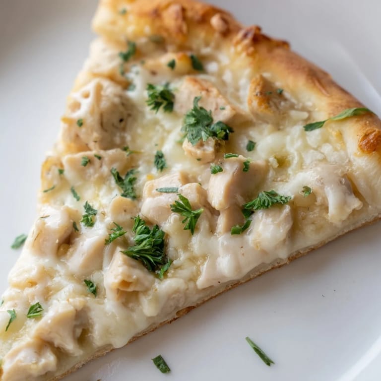 A close-up slice of Chicken Alfredo Pizza showing a crispy edge, rich Alfredo sauce, and tender chicken pieces on a wooden cutting board.  